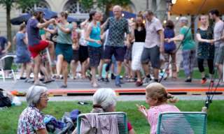 Drei Frauen sitzen auf bequemen Stühlen im Innenhof des Berner Generationenhaus, im Hintergrund tanzen viele Leute.