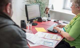 Eine Frau erklärt einem Mann etwas Computer, beide sitzen an einem Pult.