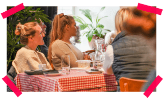 Zwei junge Frauen sitzen mit anderen Menschen an einem Tisch und lauschen einem Vortrag.
